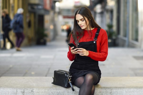 Portrait of fashionable young woman sitting on bench at pedestrian area looking at cell phone